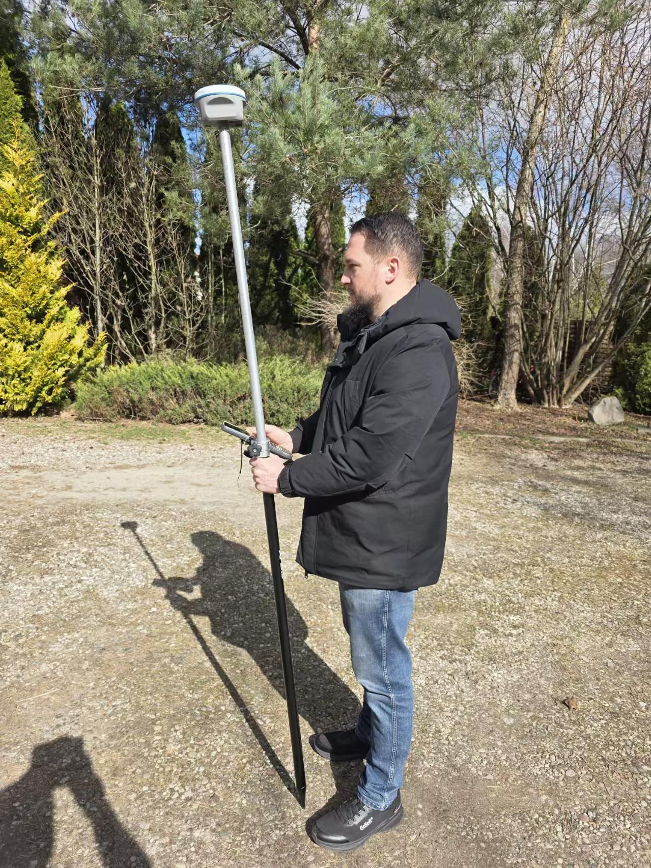 Surveyor carrying an APEKS RTK GNSS rover pole during a rural field survey in a Polish pine forest environment